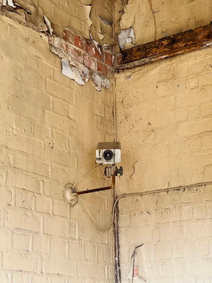 Old security camera mounted in a decaying corner with peeling paint and exposed brick.